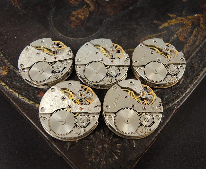 May include: Five vintage watch movements with visible gears and intricate mechanical details. The silver-toned components include small screws, wheels, and a balance wheel, with gold and red accents. The watch parts are arranged on a dark, textured surface.