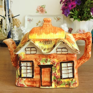 Pode incluir: Uma chaleira de cerâmica em forma de casa de campo, com telhado de colmo e janelas. A chaleira é pintada em tons de laranja, amarelo e castanho, com asa e bico. Um objeto decorativo para a cozinha.