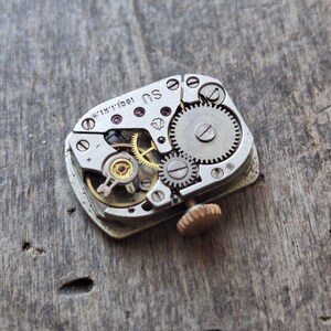 Puede incluir: Primer plano de un movimiento de reloj vintage con intrincados engranajes, tornillos y una tija de cuerda. Los componentes met&aacute;licos son plateados, con algunos engranajes dorados. El mecanismo se encuentra sobre una superficie de madera envejecida.