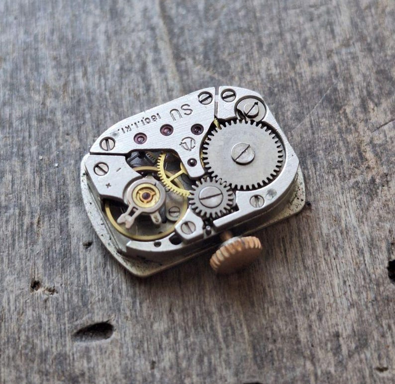 Puede incluir: Primer plano de un movimiento de reloj vintage con intrincados engranajes, tornillos y una tija de cuerda. Los componentes met&aacute;licos son plateados, con algunos engranajes dorados. El mecanismo se encuentra sobre una superficie de madera envejecida.