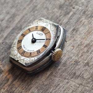 May include: Vintage Luch wristwatch with a silver case and a gold-toned face. The watch face features Roman numerals and the text "Luch" and "Made in Belarus". The watch has a small, textured crown and ornate detailing around the face.
