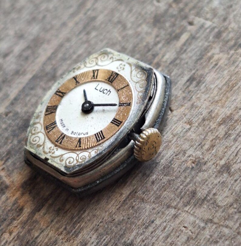 May include: Vintage Luch wristwatch with a silver case and a gold-toned face. The watch face features Roman numerals and the text "Luch" and "Made in Belarus". The watch has a small, textured crown and ornate detailing around the face.