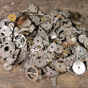 Peut inclure: Un tas de pièces de montre anciennes, comprenant des engrenages, des ressorts et des plaques, de formes et de tailles variées. Les composants sont en métal argenté et disposés sur une surface en bois. L'image met en valeur les détails complexes des mécanismes horlogers.