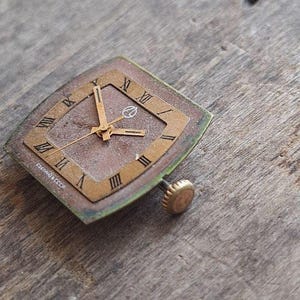 Peut inclure: Cadran de montre vintage de forme carr&eacute;e en losange. Il pr&eacute;sente des chiffres romains, des aiguilles dor&eacute;es et un petit logo. Le cadran a un cadre dor&eacute; et un cadran marron. Le texte "Сделано в СССР" est visible.