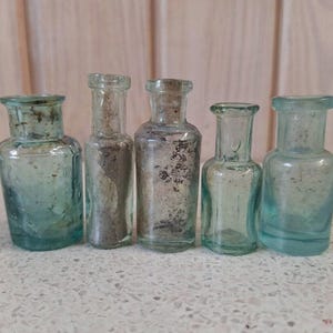 Può includere: Cinque bottiglie di vetro antiche di varie forme e dimensioni. Le bottiglie sono in tonalità di blu e trasparente, con segni di usura visibili. Sono disposte in fila, suggerendo una collezione o un'esposizione di oggetti vintage.