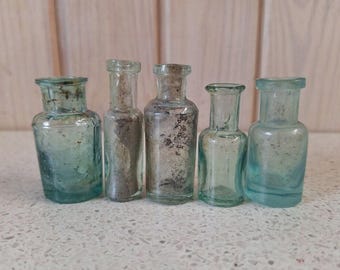 May include: A collection of five vintage glass bottles in various shapes and sizes. The bottles are in shades of blue and clear, showing signs of age. They are arranged in a row, suggesting a display of antique items.