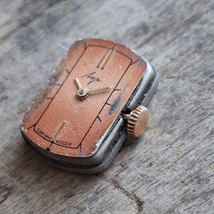 Puede incluir: Reloj de pulsera rectangular vintage con esfera color cobre y agujas doradas. El reloj tiene una caja plateada y una corona dorada. La esfera tiene marcas negras y la palabra "Луч" en dorado. El reloj est&aacute; marcado "Сделано в СССР".
