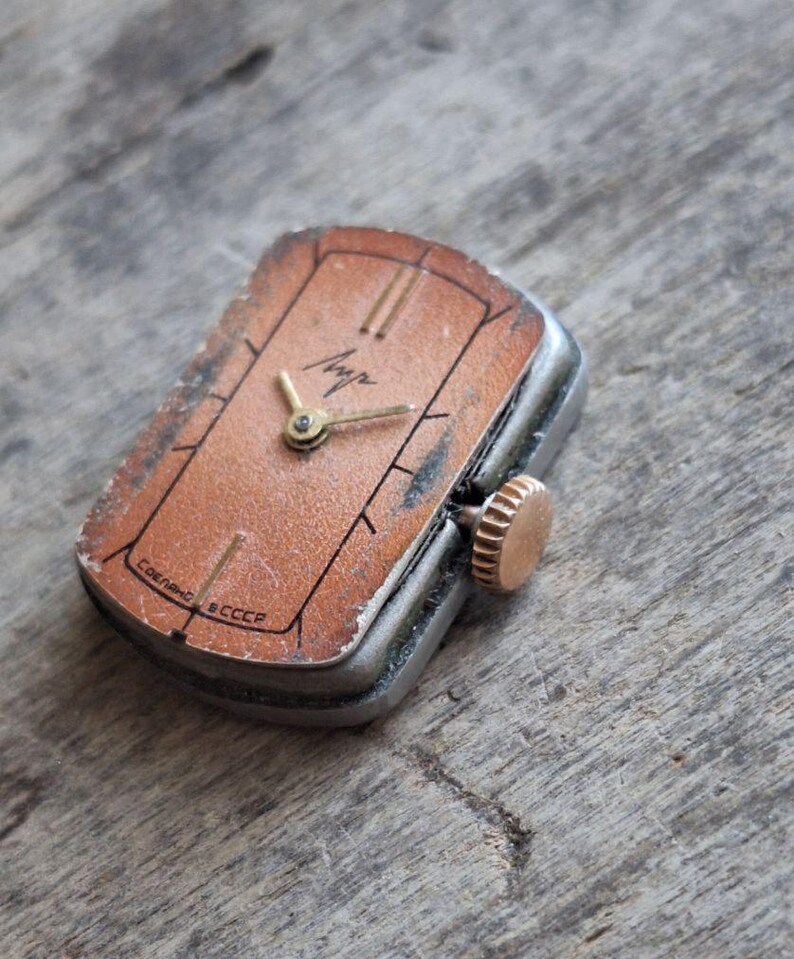 Puede incluir: Reloj de pulsera rectangular vintage con esfera color cobre y agujas doradas. El reloj tiene una caja plateada y una corona dorada. La esfera tiene marcas negras y la palabra "Луч" en dorado. El reloj est&aacute; marcado "Сделано в СССР".