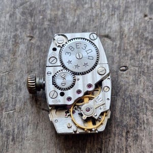 Peut inclure: Gros plan d'un mouvement de montre vintage avec des engrenages, des vis et une tige de remontoir complexes. Les composants m&eacute;talliques sont argent&eacute;s et dor&eacute;s, avec le mot "ЗВЕЗДА" visible sur un engrenage. Le mouvement est pos&eacute; sur une surface en bois vieilli.