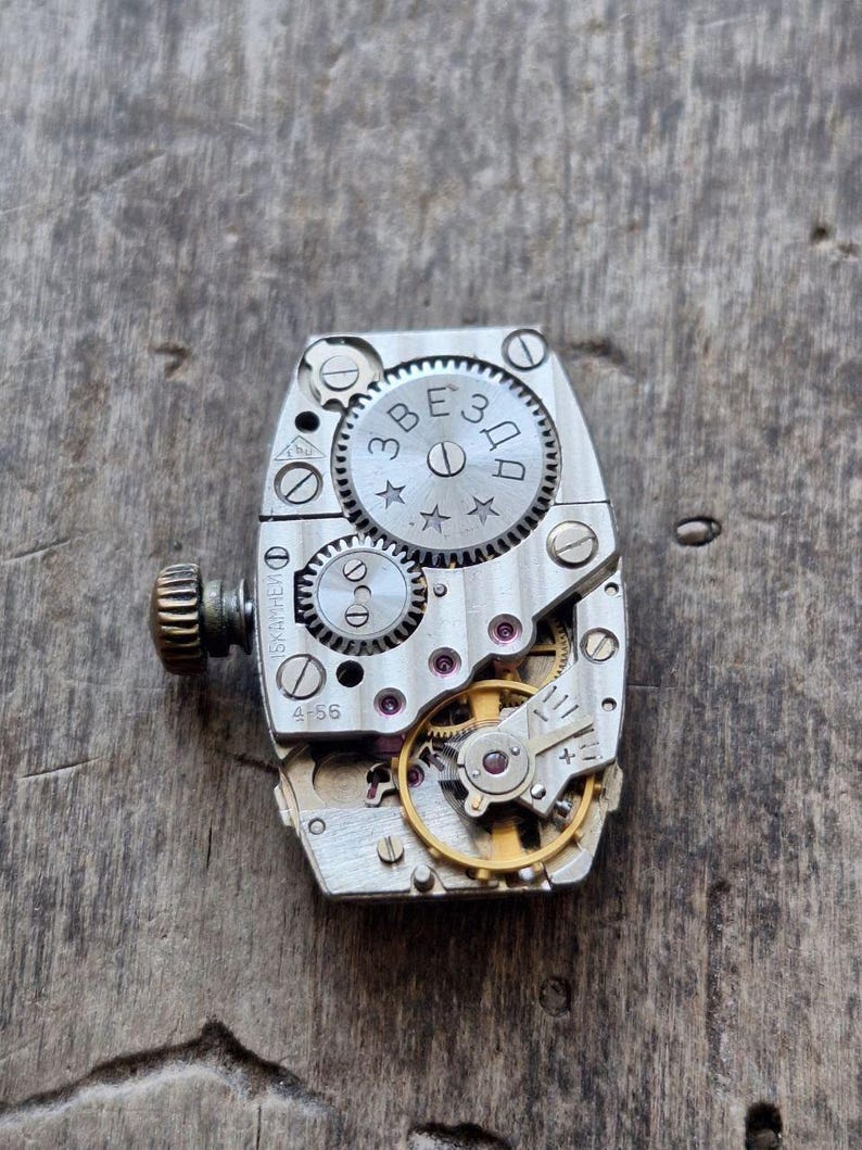 Peut inclure: Gros plan d'un mouvement de montre vintage avec des engrenages, des vis et une tige de remontoir complexes. Les composants m&eacute;talliques sont argent&eacute;s et dor&eacute;s, avec le mot "ЗВЕЗДА" visible sur un engrenage. Le mouvement est pos&eacute; sur une surface en bois vieilli.