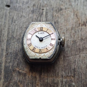 May include: Vintage Luch wristwatch with a silver-tone case and a cream-colored face. The watch features gold Roman numerals and hands, with the text "Luch" and "Made in Belarus" visible. The watch has a small crown and is set against a wooden surface.