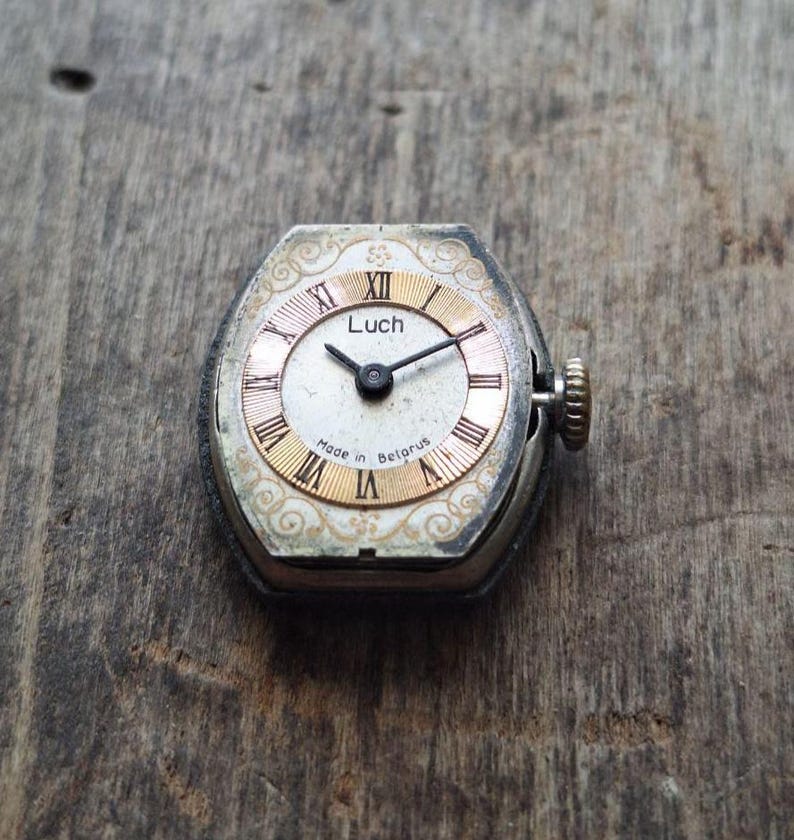 May include: Vintage Luch wristwatch with a silver-tone case and a cream-colored face. The watch features gold Roman numerals and hands, with the text "Luch" and "Made in Belarus" visible. The watch has a small crown and is set against a wooden surface.