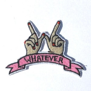 May include: Embroidered patch featuring two hands forming the peace sign, with red nail polish. A pink banner below reads "WHATEVER" in white letters. The patch has a white border and is likely for clothing or accessories.