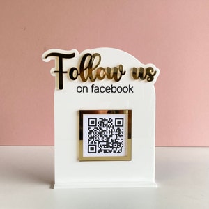Könnte beinhalten: Weißes Schild mit goldener Aufschrift "Follow us on facebook" und einem goldenen Rahmen mit einem schwarz-weißen QR-Code.