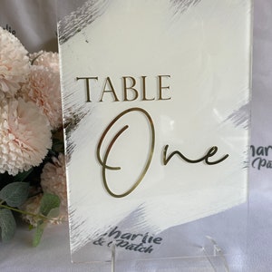 May include: White acrylic table number sign with gold lettering. The sign reads "TABLE One" and has a white brushstroke design. The sign is on a clear acrylic stand.