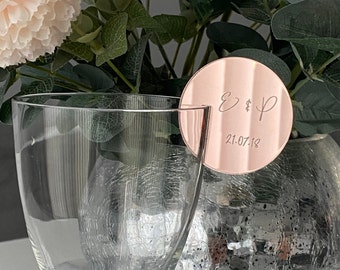 Op de afbeelding: Een ronde plaatskaart van rosé goud met de initialen "E & P" en de datum "21.07.18" erop gegraveerd. De plaatskaart ligt op een helder glas.