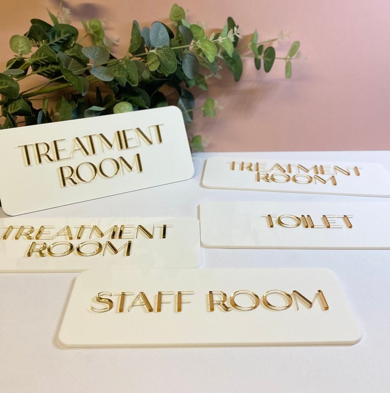 May include: Five white rectangular signs with gold lettering. The signs read "Treatment Room", "Treatment Room", "Toilet", "Staff Room", and "Treatment Room".