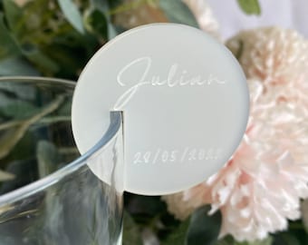 Op de afbeelding: Een witte, ronde drankbedel met de naam "Julian" en de datum "28/05/2023" gegraveerd. De bedel is ontworpen om aan de rand van een glas te hangen. De achtergrond bevat zachte roze en groene bloemstukken.