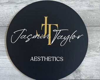 May include: A black, circular sign with the text "Jasmin Taylor" in white script and gold initials. The word "AESTHETICS" is printed in white block letters below. The sign is set against a gray wood-grain background.