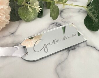 May include: A silver mirror tag with the name "Gemma" engraved on it. The tag is attached to a white ribbon.