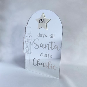 May include: An arched mirrored acrylic sign featuring an etched nutcracker figure on the left side. The text reads "days till Santa visits Charlie" with a white star-shaped attachment at the top displaying the handwritten number 131.