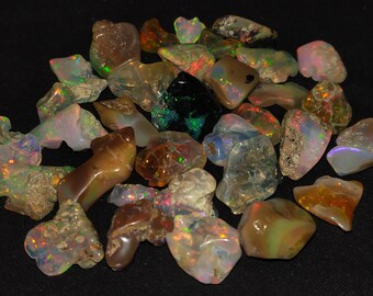 Puede incluir: Una colección de piedras preciosas de ópalo natural en bruto en varios tonos de marrón, blanco y verde. Los ópalos tienen un brillo iridiscente y se muestran sobre un fondo negro.