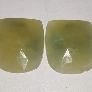 May include: Two faceted, cushion-cut gemstones. The gems are a pale yellow-green color with internal inclusions and a polished surface. The facets create a sparkling effect, and the stones have rounded corners.