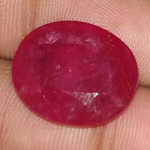 May include: A deep red oval gemstone with a smooth, polished surface. The stone has a slight sparkle and is set in a clear, faceted setting.