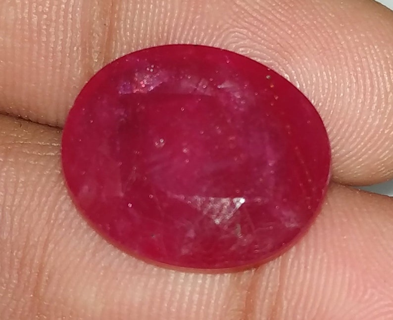 May include: A deep red oval gemstone with a smooth, polished surface. The stone has a slight sparkle and is set in a clear, faceted setting.