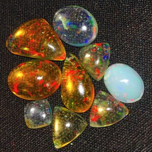 May include: A collection of eight opal gemstones in various shapes and sizes. The opals are a mix of clear, white, yellow, and orange with flashes of iridescent color.