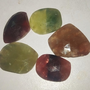 May include: An assortment of faceted gemstones in various colors, including shades of red, green, and brown. The gemstones are irregularly shaped and appear to be polished. The arrangement suggests a collection of loose stones.