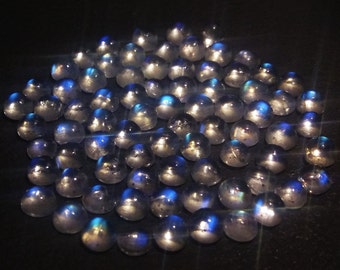 Op de afbeelding: Een verzameling gepolijste, iriserende maansteen cabochons. De stenen zijn grotendeels rond met een glad, gewelfd oppervlak. Elke steen reflecteert een spel van blauw en zilver licht, waardoor een glinsterend effect ontstaat. De stenen zijn dicht bij elkaar op een donkere ondergrond geplaatst.