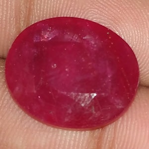 May include: A deep red oval gemstone with a faceted cut. The gemstone has a slightly cloudy appearance with visible inclusions.