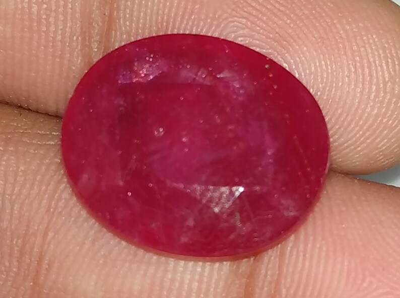 May include: A deep red oval gemstone with a faceted cut. The gemstone has a slightly cloudy appearance with visible inclusions.