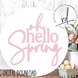 Puede incluir: Un letrero redondo de madera blanca con el texto rosa "Oh Hello Spring". El letrero está sobre una superficie de madera con una cesta de huevos y una planta en maceta con flores rosas.