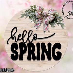 May include: A round wooden sign with a white background and the text "hello spring" in black. The sign is decorated with a floral bow with pink and white flowers.