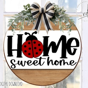 May include: A round wooden sign with a white background and the text "Home sweet home" in black script. A red ladybug with black spots is centered above the text. A green leafy wreath with a black and white bow is above the sign.
