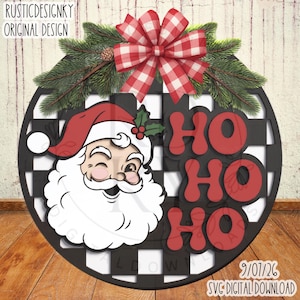 May include: A round wooden sign featuring a black and white buffalo check pattern, a winking Santa Claus illustration, and the red text HO HO HO. The sign is decorated with a red and white gingham bow and green pine branches with a small pinecone.