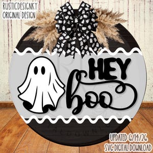 May include: A round wooden door sign featuring a black and white ghost pattern bow, dried pampas grass, and a white center panel with a black ghost illustration and the text HEY BOO. The sign has black horizontal wood grain sections at the top and bottom.