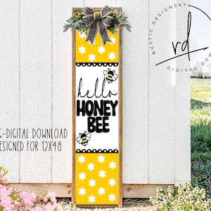 May include: A wooden sign with a yellow and white background featuring a bee and the text "hello honey bee". The sign is decorated with a black and white bow and a floral wreath.