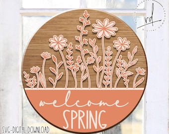 May include: A round wooden sign with a peach background and a white outline of flowers and stems. The sign says "Welcome Spring" in white text.