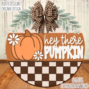May include: A round wooden door sign featuring a brown and white checkered pattern at the bottom, an orange pumpkin with two white daisies, and the text "hey there PUMPKIN" in white. A brown and white gingham bow sits at the top with green leaves.