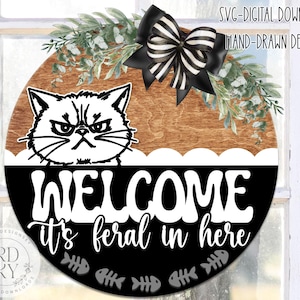 Può includere: Un cartello di benvenuto rotondo in legno con un'illustrazione di gatto in bianco e nero e la scritta "WELCOME it's feral in here." Il cartello è decorato con un fiocco a righe bianche e nere e vegetazione. Download digitale.