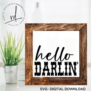 May include: A wooden framed sign with the text "hello darlin'" in black lettering. The sign is sitting on a white surface with a green plant in a metal pot.