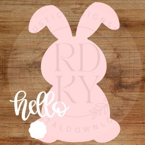 May include: A pink bunny-shaped cutout with a white fluffy tail and the word "hello" in cursive. The bunny has long ears and is set against a brown wood grain background. A decorative item for Easter.