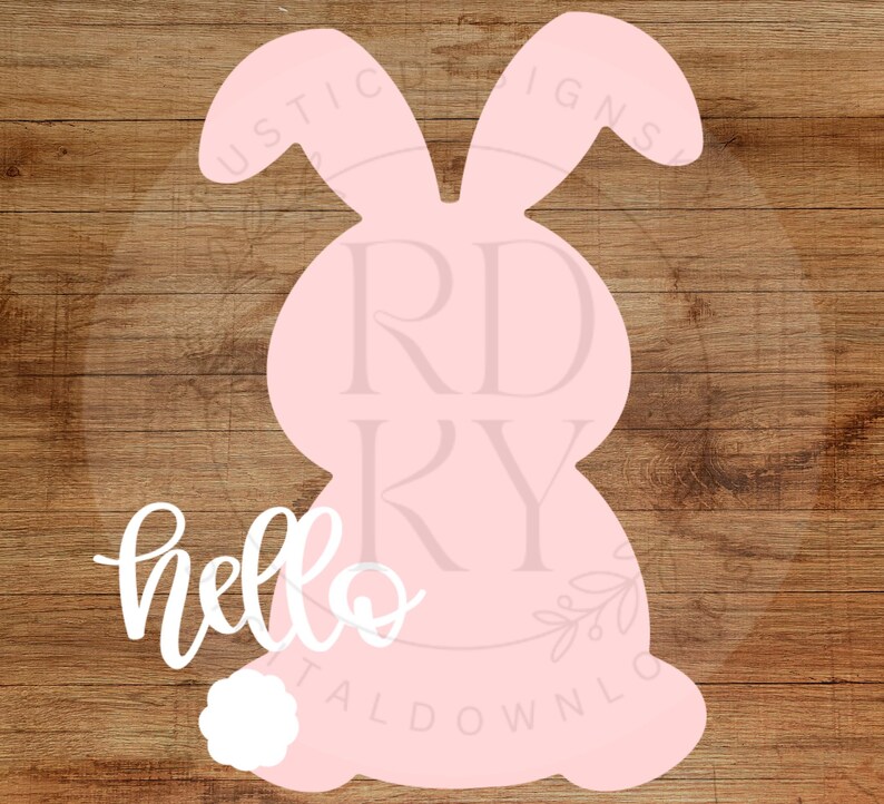 May include: A pink bunny-shaped cutout with a white fluffy tail and the word "hello" in cursive. The bunny has long ears and is set against a brown wood grain background. A decorative item for Easter.