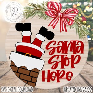 Op de afbeelding: Een ronde houten kerstbord met een ontwerp van de kerstman, met zijn benen uit een schoorsteen. Het bord leest "Santa Stop Here" in rood script. Een rood-witte gestreepte strik en dennentakken staan bovenaan.