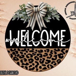 May include: A round wooden sign with a black background and a white "WELCOME" inscription. The top half of the sign features a black and white striped bow with green leaves. The bottom half of the sign features a brown and black leopard print pattern.