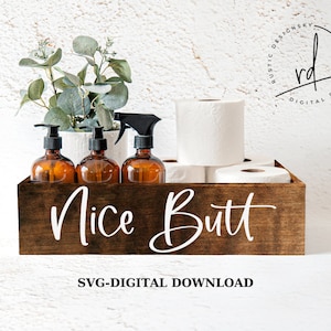 May include: A dark brown wooden bathroom organizer with the words "Nice Butt" in white script. The organizer holds three amber glass bottles with black pumps and a spray bottle, along with rolls of toilet paper and a small plant.