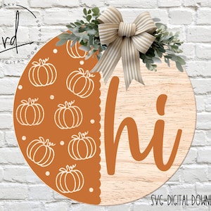 May include: A round wooden sign with a brown background and the word "hi" written in orange. The left side of the sign is decorated with a pattern of white pumpkins on an orange background.
